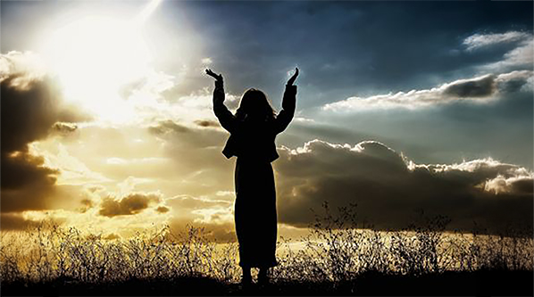 Woman in silhouette with hands up praying to God against a dramatic sky.