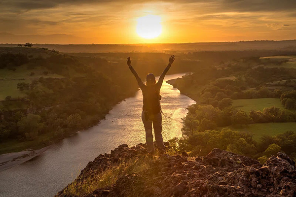 A hiker on top of a hill facing a sunset with their arms up.