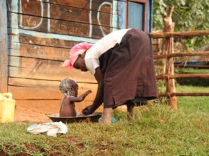Kenyan woman bathing her child outside in the yard of her home.