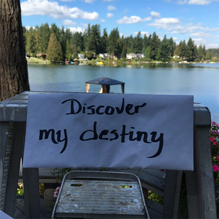 Hand written sign that says discover my destiny, outside with a lake scene behind it.